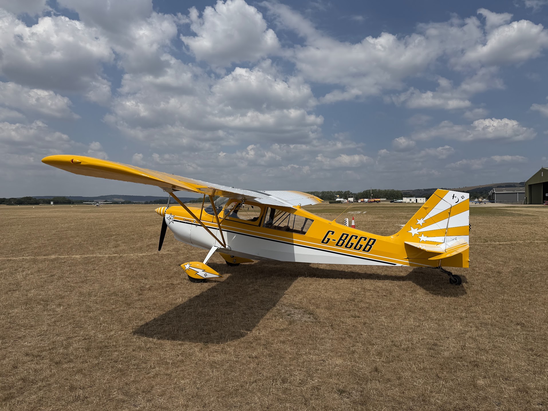 Yellow and white Citabria G-BGGB parked on a dry grass airfield beneath large clouds