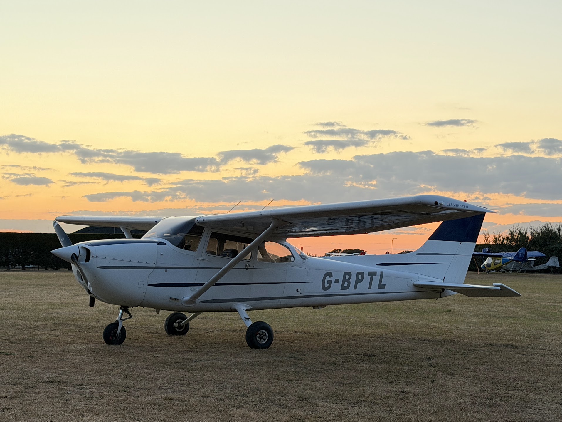 White and navy Cessna 172 G-BPTL parked on a grass airfield at sunset